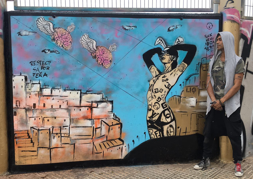 Artist White Rabbit standing beside a completed street art mural in Ibiza